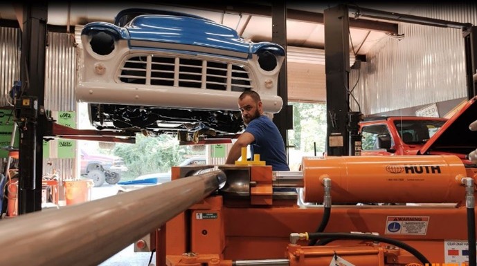 Un hombre con una camisa azul dobla una tubería automotriz gris con una máquina dobladora de tubos Huth-Ben Pearson naranja. Arriba del hombre, el frente de un auto clásico azul y blanco está en un elevador dentro de un taller de automóviles.