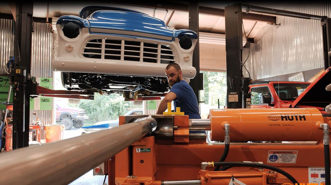 A man in a blue shirt bends a grey automotive pipe using an orange Huth-Ben Pearson tubing bender machine. Above the man, a blue and white classic car front is on a lift inside the automotive shop.